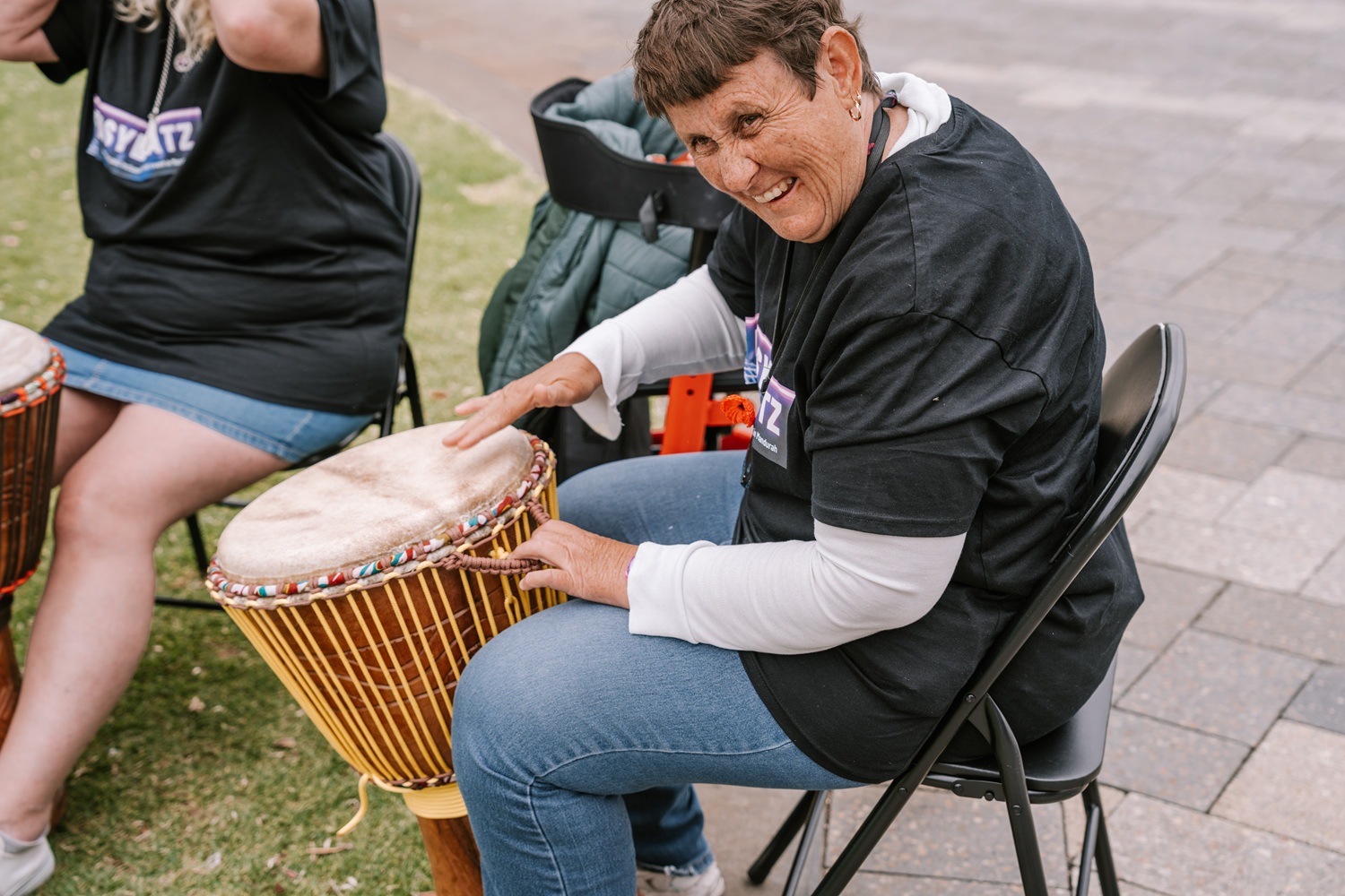 A smiling woman seated on a chair plays a hand drum during a group drumming activity.